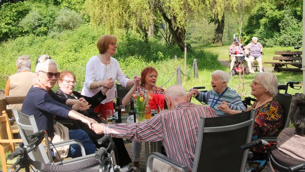 Zu sehen ist eine Gruppe von Bewohnerinnen und Bewohnern, die gemeinsam an einem Tisch im Garten sitzen und Hand in Hand schunkeln. 